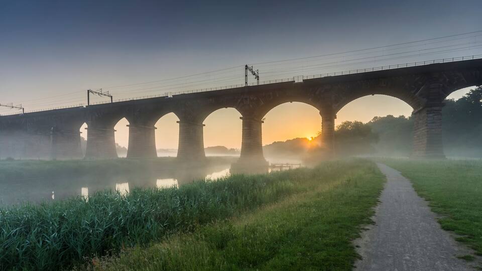 A stone bridge with multiple arches over a calm river, surrounded by greenery and a misty atmosphere.