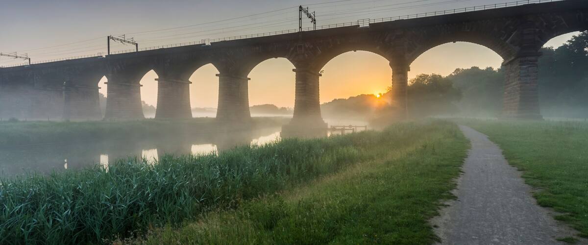 A stone bridge with multiple arches over a calm river, surrounded by greenery and a misty atmosphere.