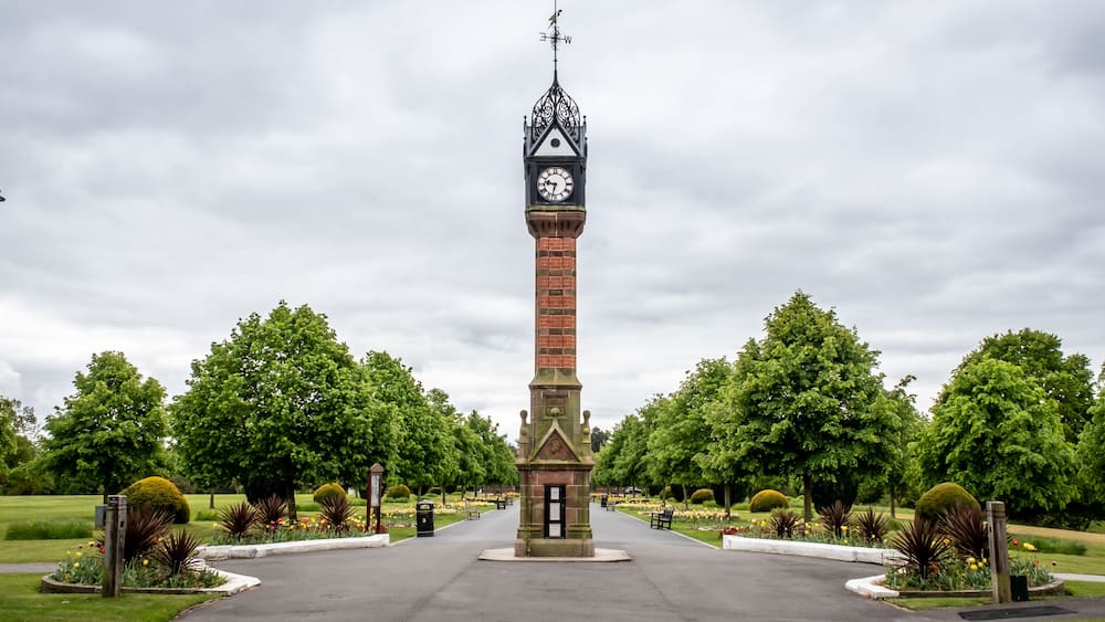 Une haute tour en briques, surmontée d’une horloge et d’une girouette, entourée d’un parc bien entretenu avec des arbres et des parterres de fleurs.