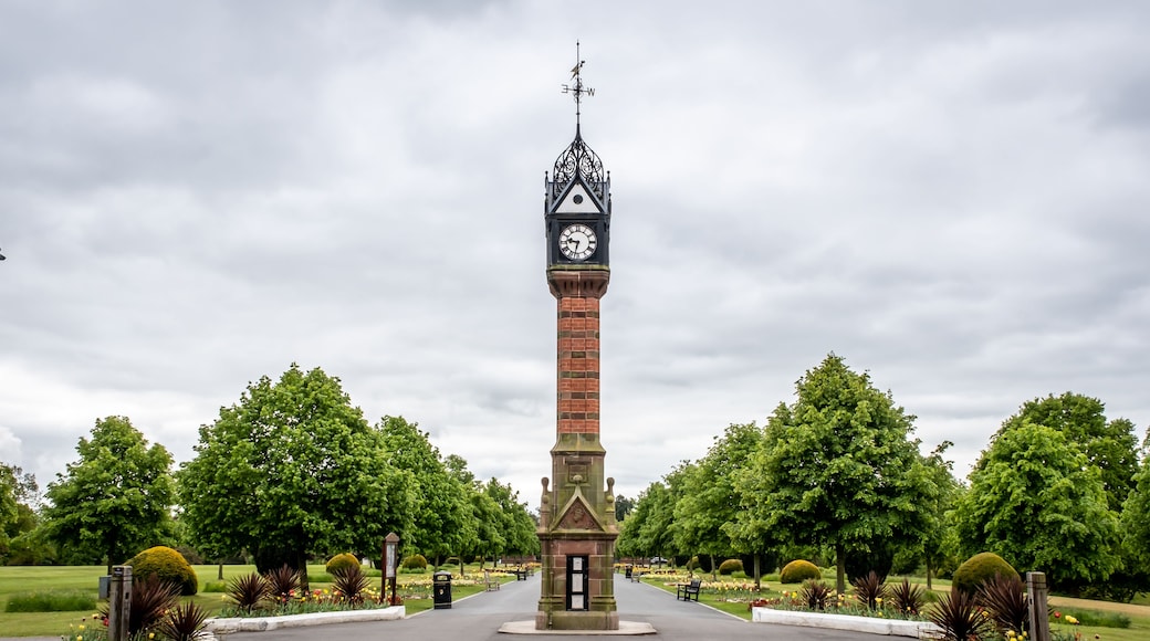 A tall brick tower with a clock and a weather vane, surrounded by a well-maintained park with trees and flower beds.