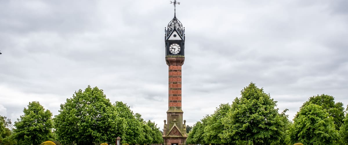 Une haute tour en briques, surmontée d’une horloge et d’une girouette, entourée d’un parc bien entretenu avec des arbres et des parterres de fleurs.