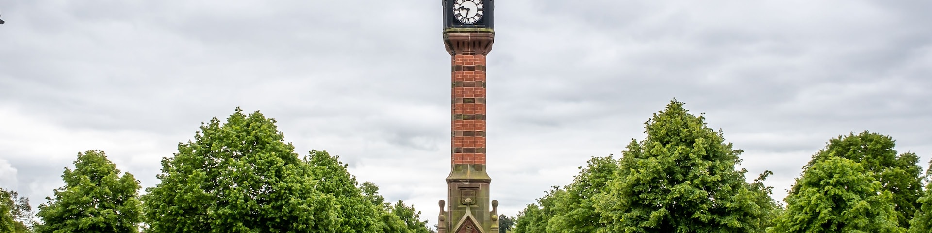 Una alta torre de ladrillo con un reloj y una veleta, rodeada por un parque bien cuidado con árboles y parterres de flores.