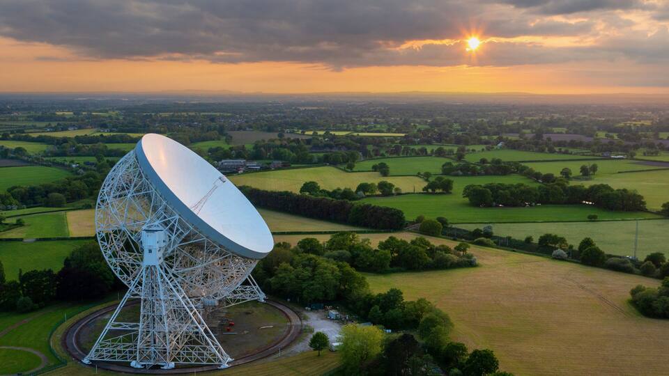 A large satellite dish in a rural landscape with fields and a sunset in the background.