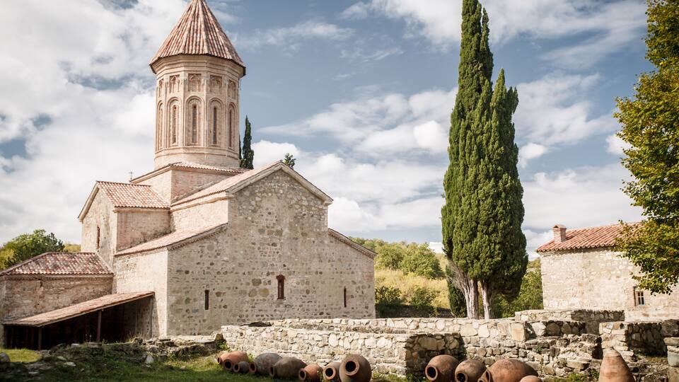 A stone church with a bell tower, surrounded by ancient pottery and cypress trees.