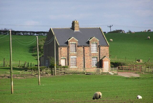 A brick house with a grey roof, surrounded by a green field and a wooden fence.