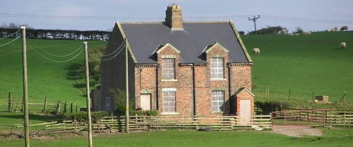 A brick house with a grey roof, surrounded by a green field and a wooden fence.