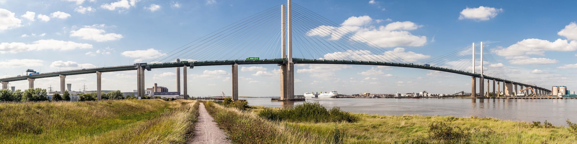 A large cable-stayed bridge spanning a river with a clear sky and industrial buildings in the background.