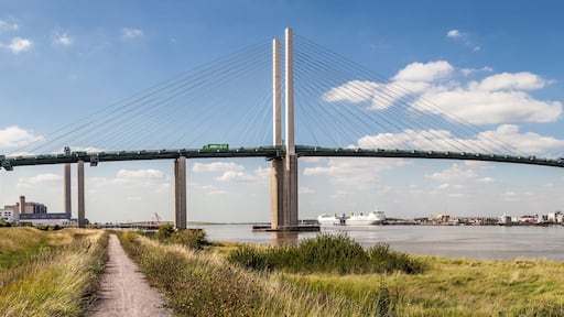 Een grote hangbrug over een rivier, met een heldere lucht en fabrieksgebouwen op de achtergrond.
