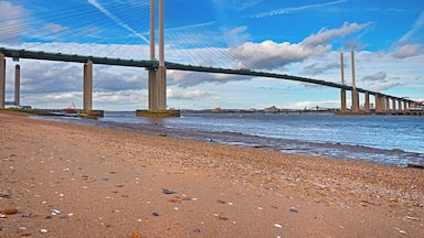 Uma grande ponte suspensa sobre um rio, com uma praia de pedras em primeiro plano.