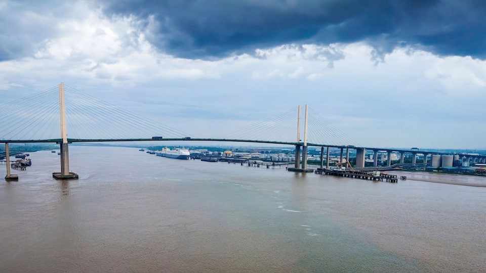 A large suspension bridge over a river with industrial buildings in the background.