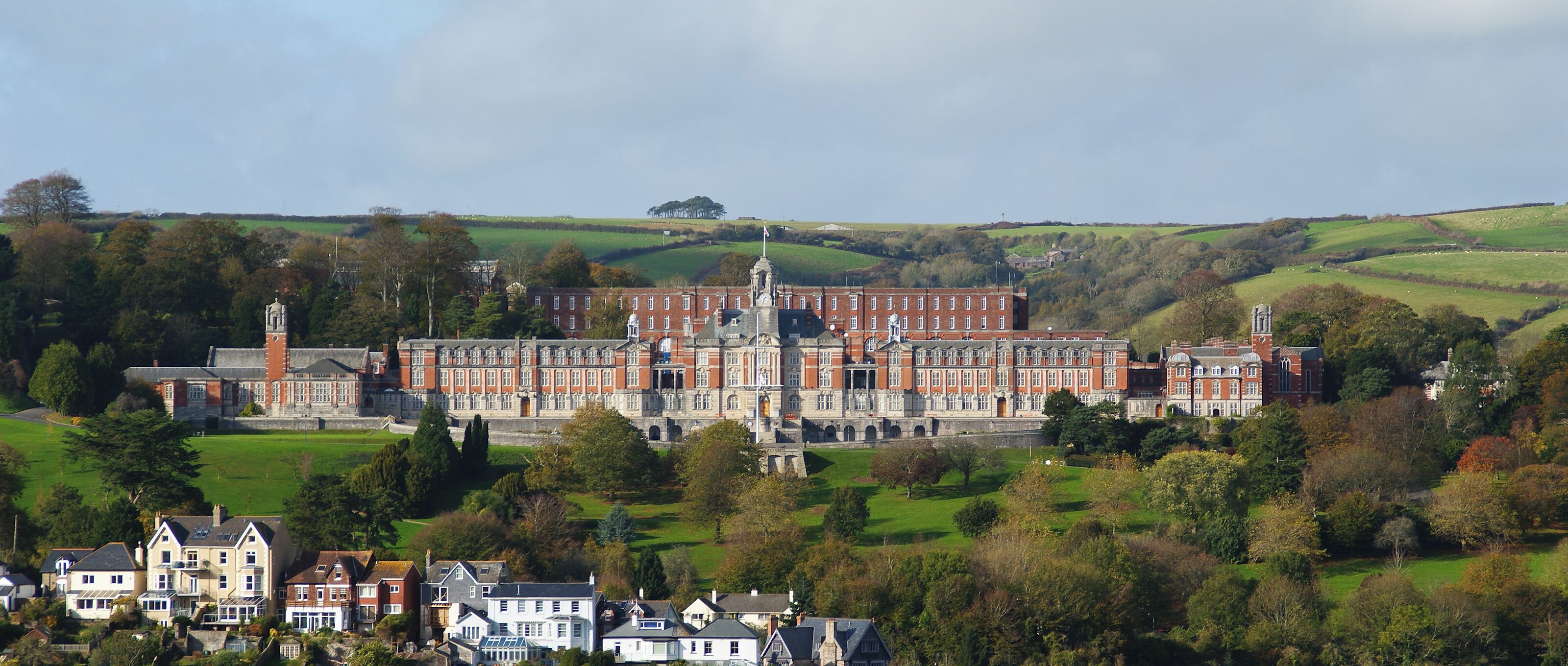 A large red brick building with multiple towers, surrounded by green fields and trees.