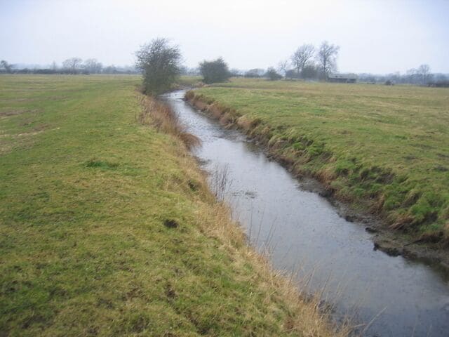A narrow water channel in a grassy field with trees in the background.
