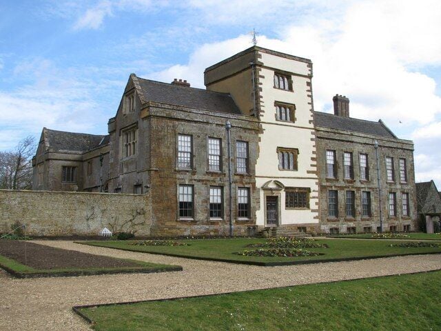 A large, historic building with a symmetrical facade, a central tower, and a well-maintained garden with geometric flower beds.