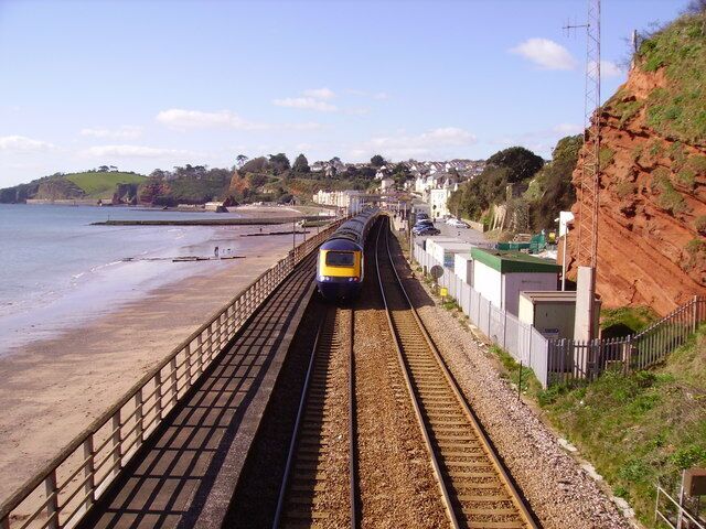 A train on tracks parallel to a beach with houses and cliffs in the background.