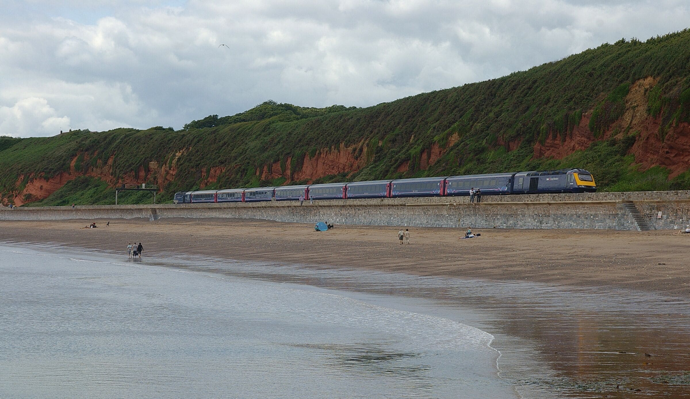 Un tren sobre rieles, paralelo a una playa donde gente camina y se sienta sobre la arena.