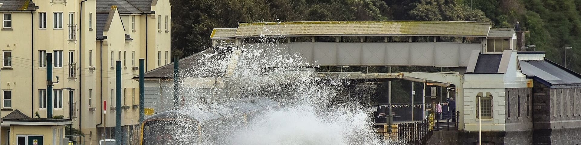 A large wave crashing over a sea wall near a coastal town with buildings in the background.