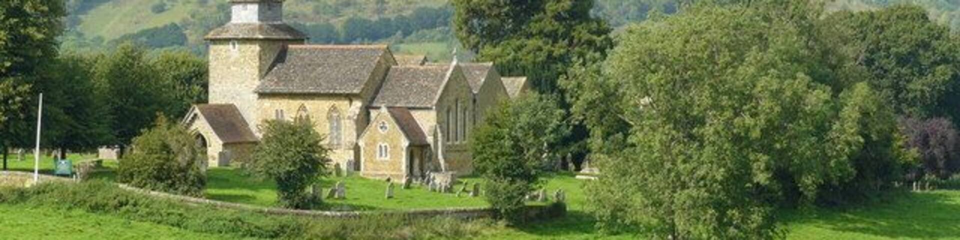 A church surrounded by lush green fields and trees, with a hill in the background.