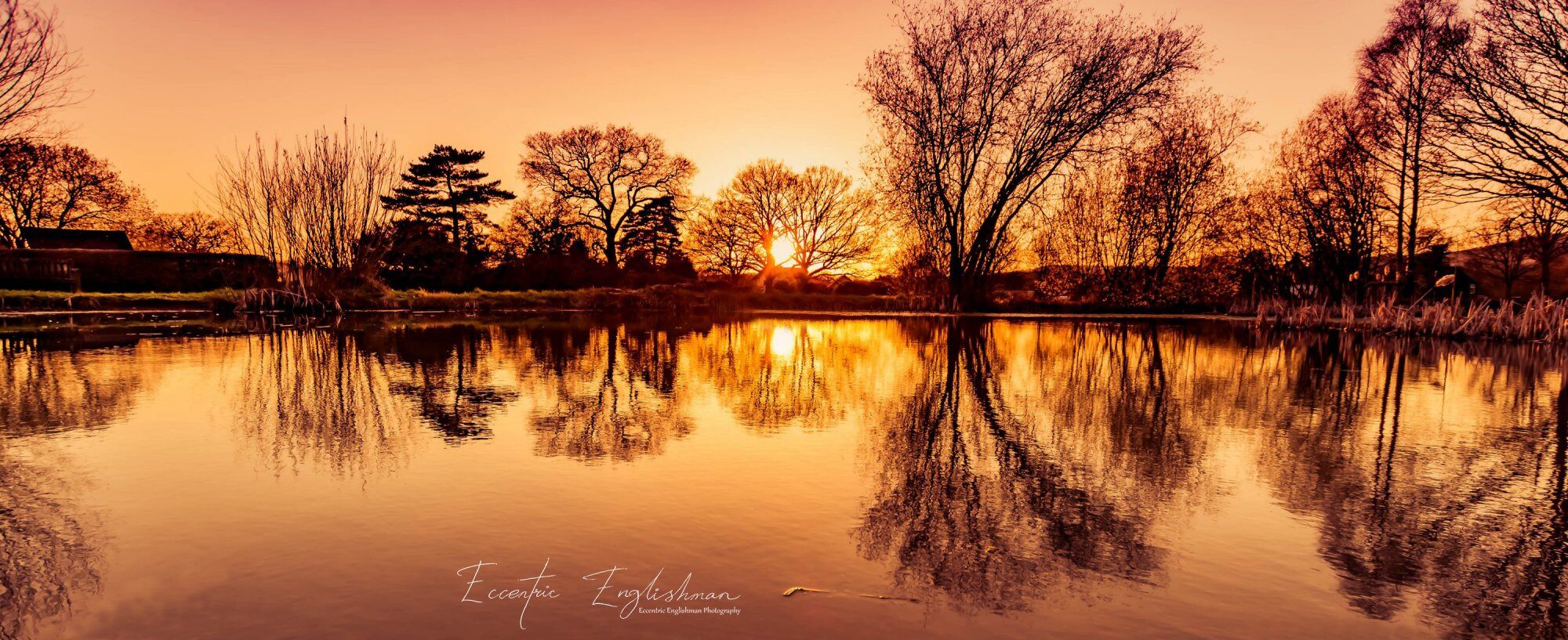 A calm lake at sunset with reflections of trees and a building.
