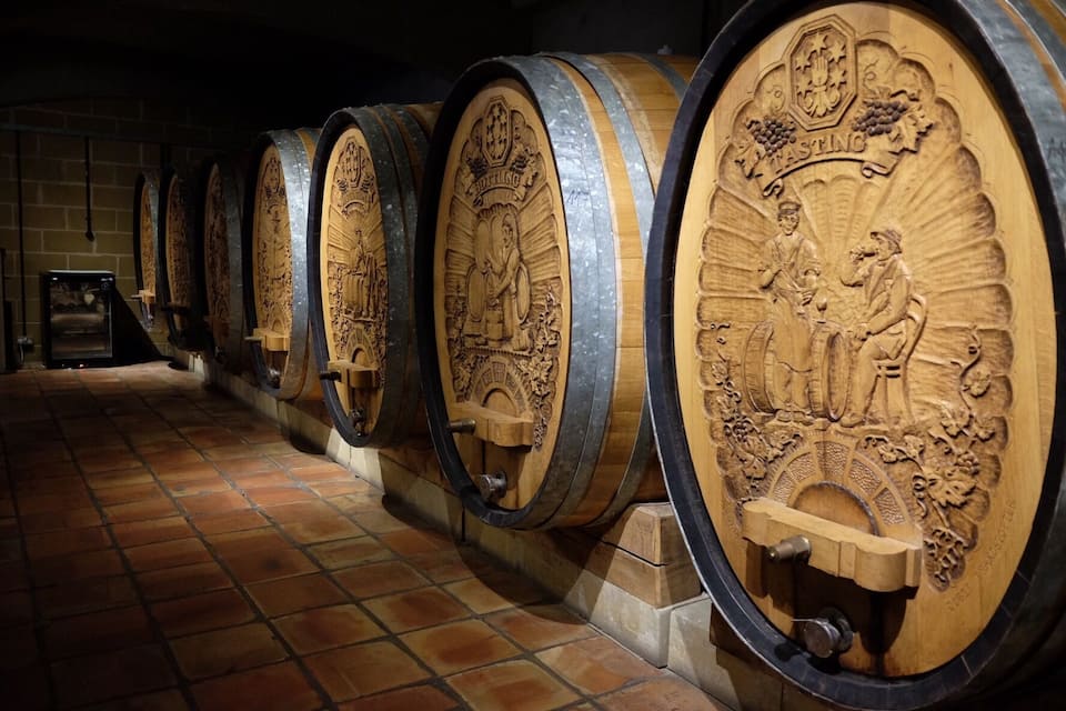 A row of wooden wine barrels with intricate carvings, a tiled floor, and a brick wall in the background.