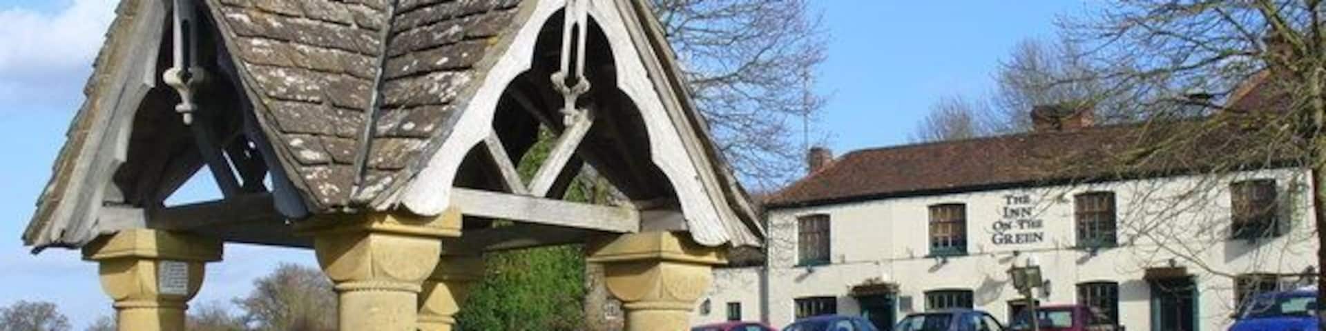A stone gazebo with a bench, a white building labeled 'The Inn of the Green Dragon' in the background, and parked cars.