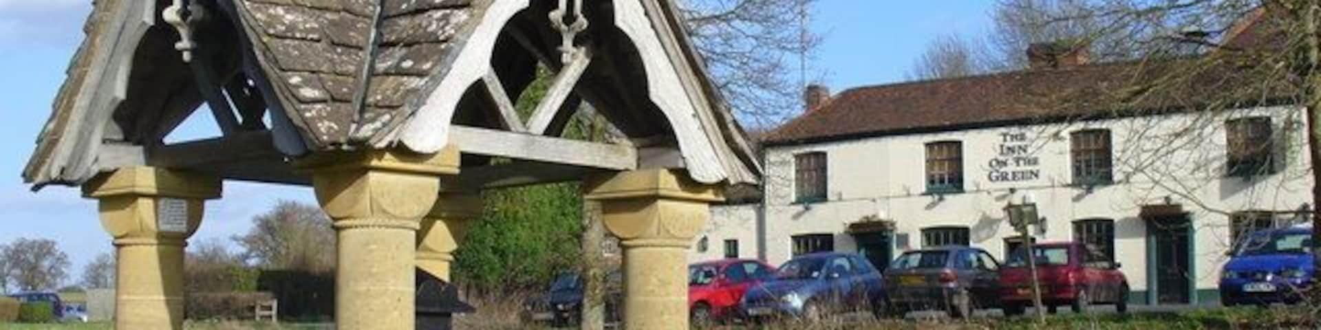Un pabellón de piedra con un banco, un edificio blanco con la inscripción 'The Inn of the Green Dragon' y coches aparcados al fondo.