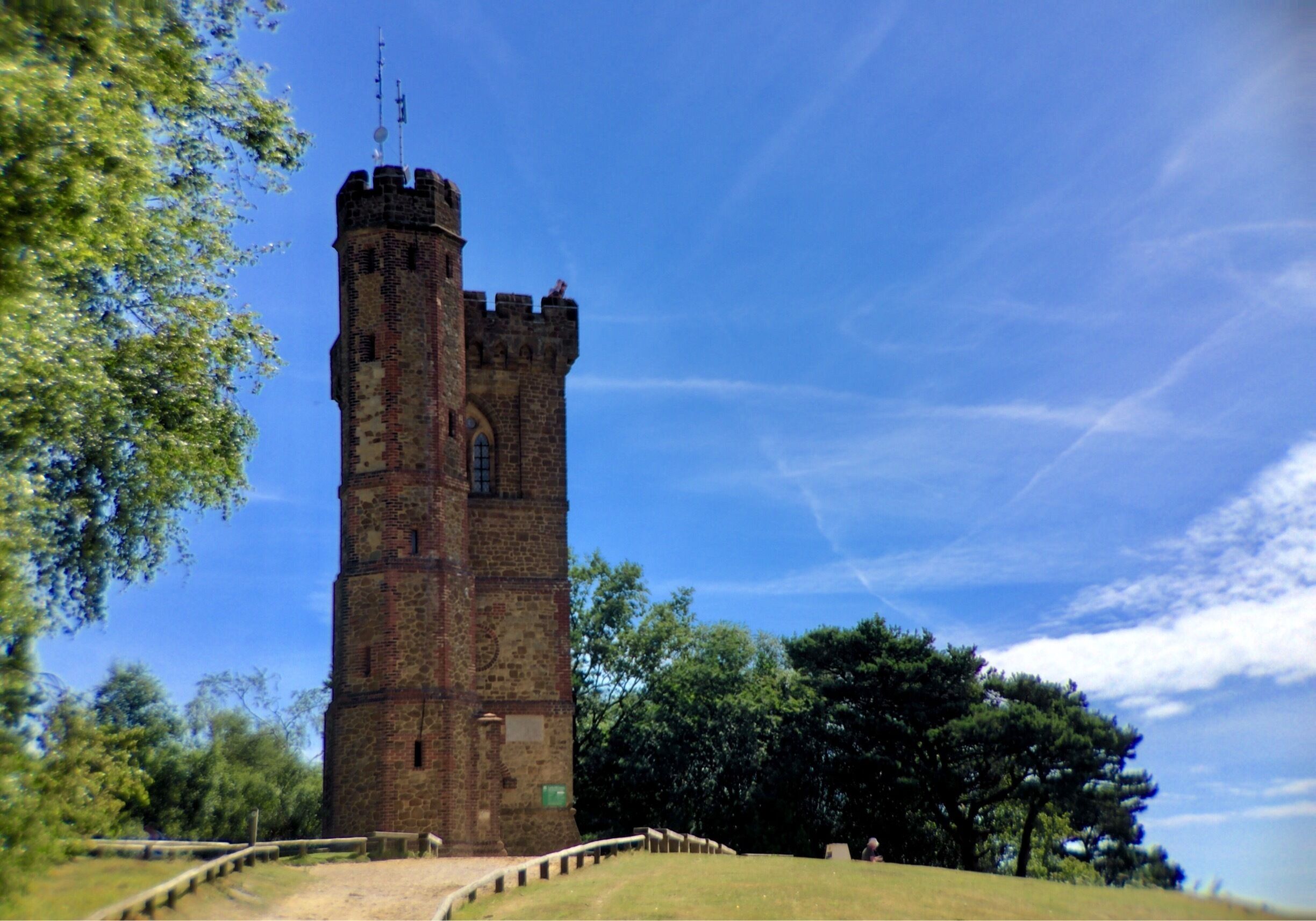 A tall stone tower with a weather vane on top, surrounded by trees and a clear sky.