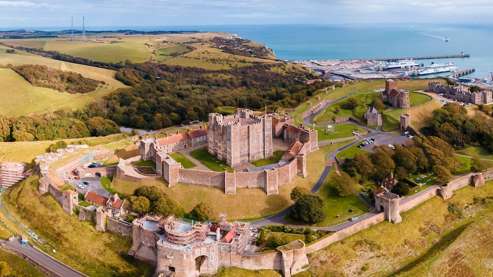 An aerial view of a historic castle surrounded by a fortified wall, with a port and a lighthouse in the background.