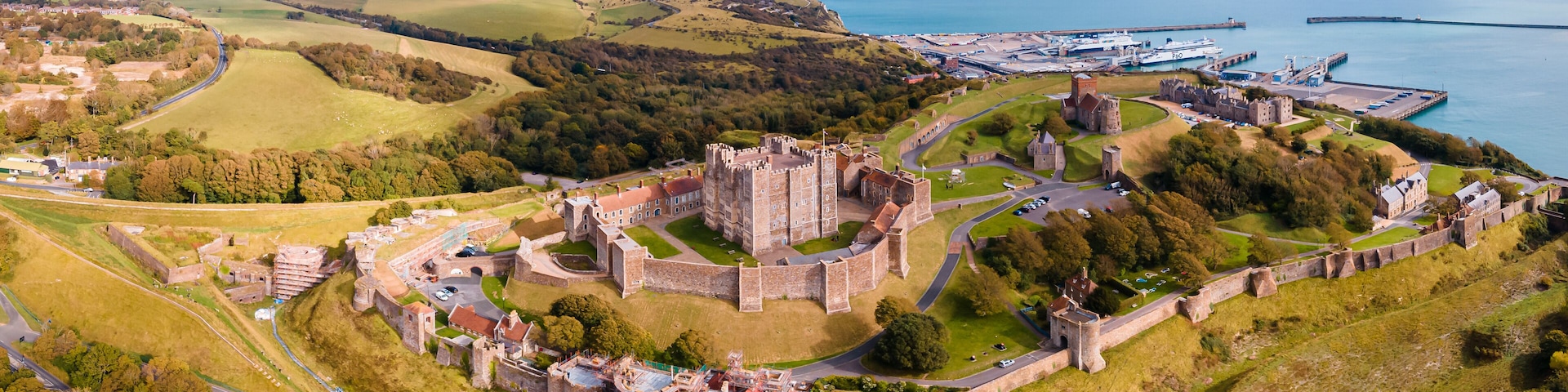 An aerial view of a historic castle surrounded by a fortified wall, with a port and a lighthouse in the background.