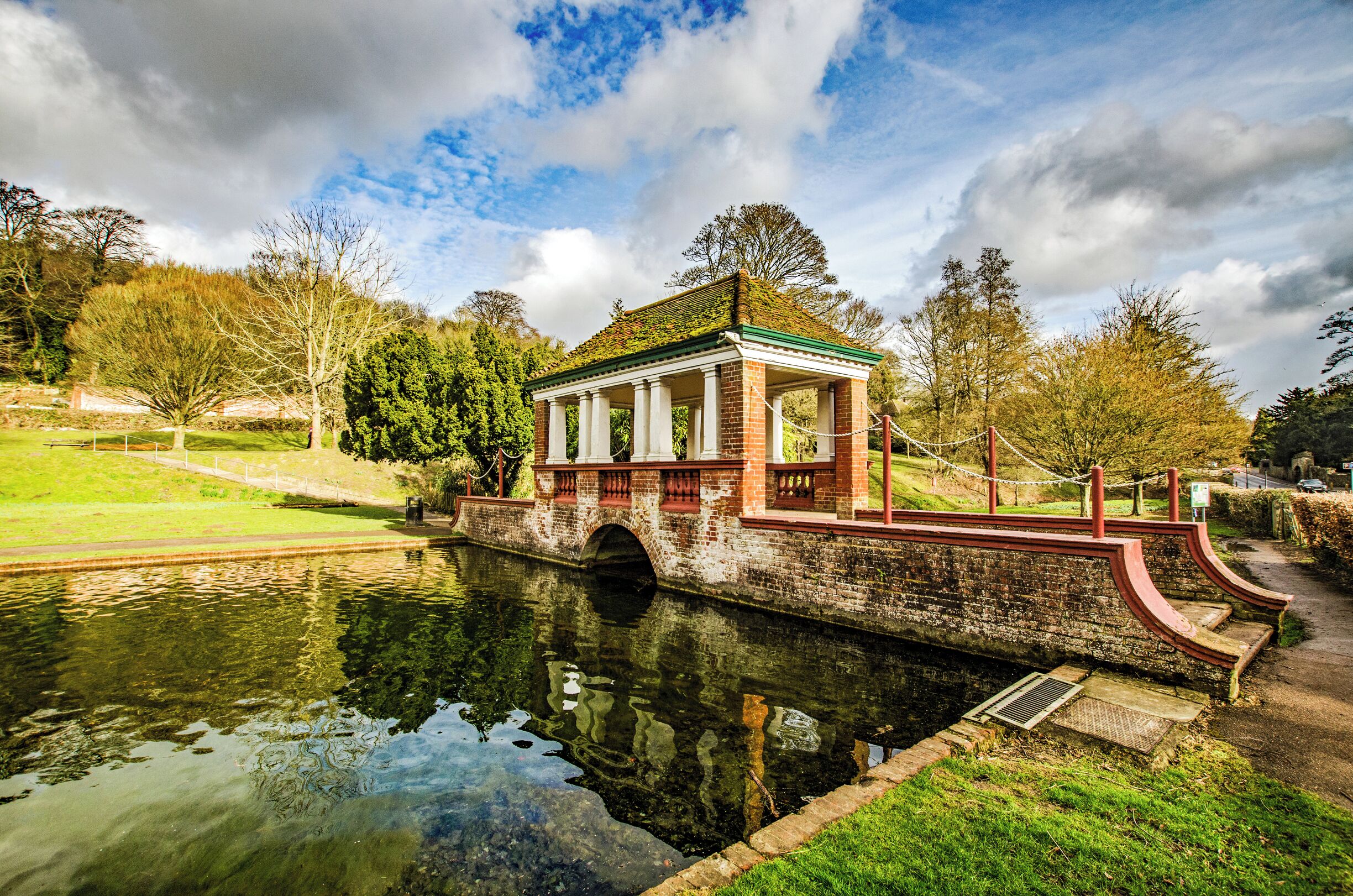 A brick pavilion with a green roof by a calm river, surrounded by trees and grassy areas.