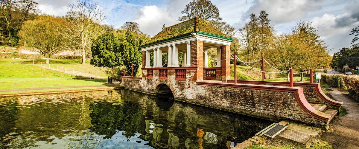 A brick pavilion with a green roof by a calm river, surrounded by trees and grassy areas.
