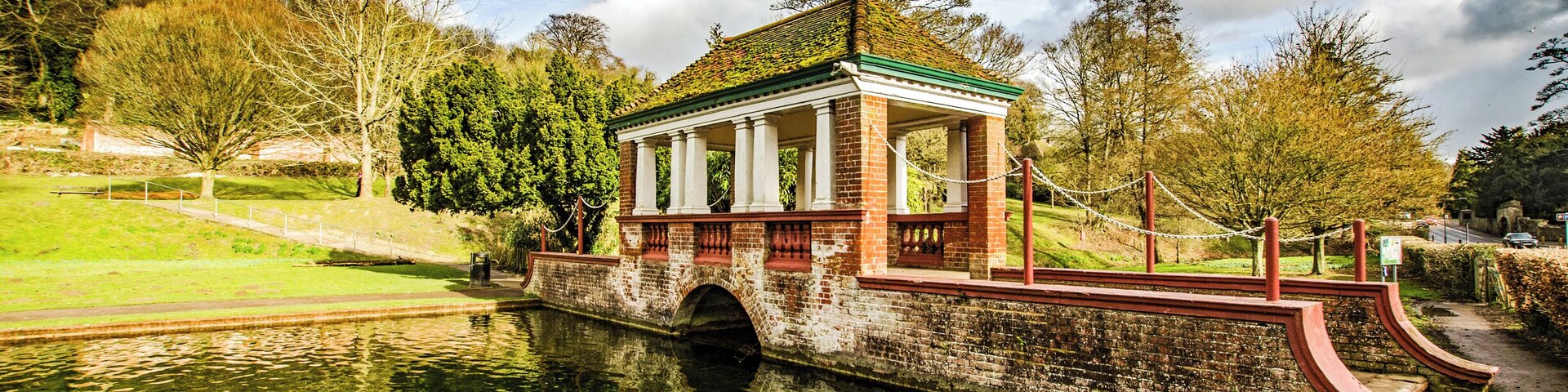 A brick pavilion with a green roof by a calm river, surrounded by trees and grassy areas.