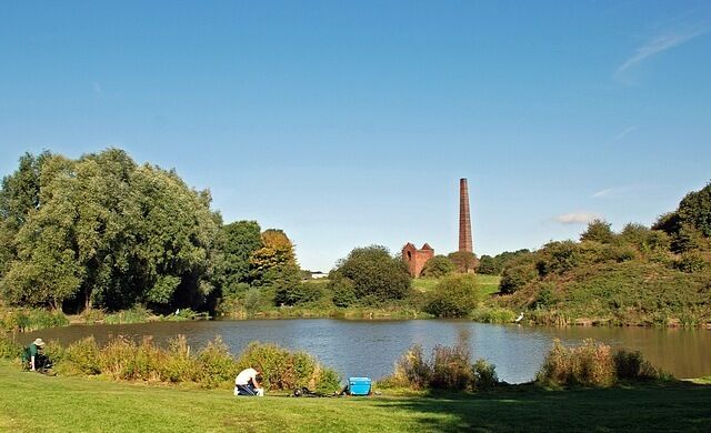 A lake with a grassy bank, trees, and a tall chimney in the background.