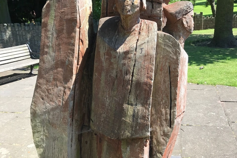 A large wooden sculpture of a figure with a staff, set in a park with a stone bench and trees in the background.