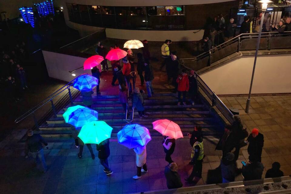 Escena nocturna con gente caminando por unas escaleras, algunos con paraguas.