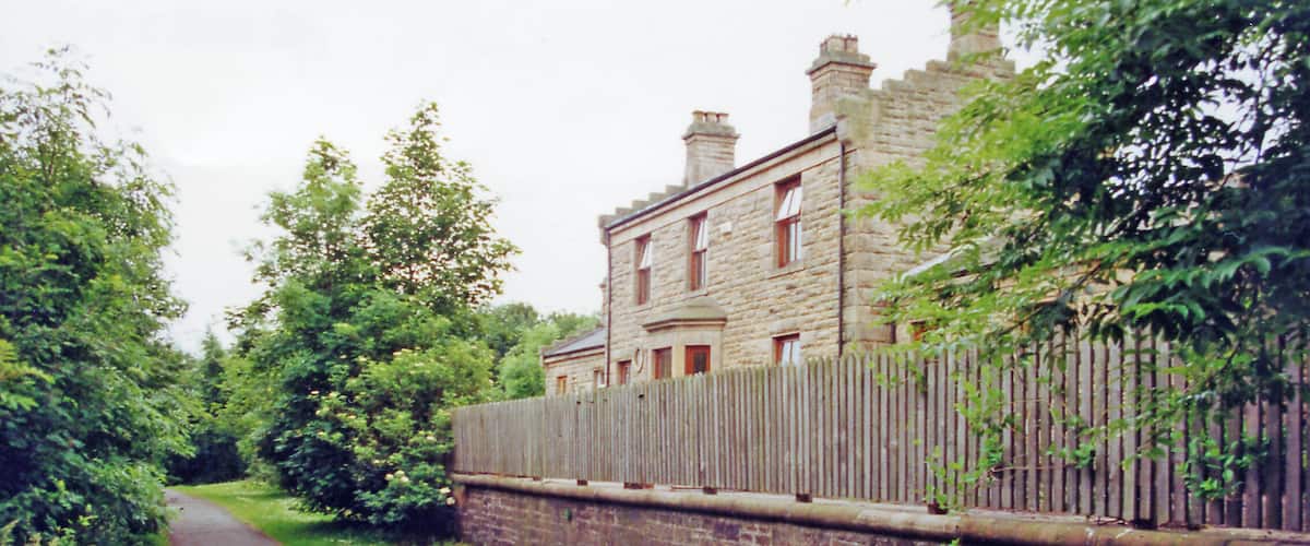 A narrow paved path flanked by lush greenery and a stone wall leading to a historic stone building with multiple chimneys.