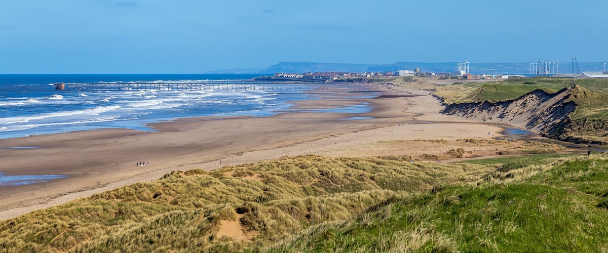 A coastal landscape with a sandy beach, grassy dunes, a pier extending into the sea, and wind turbines in the distance.