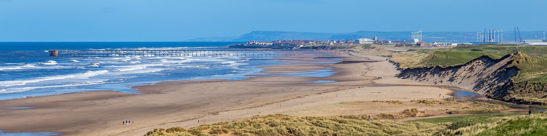 A coastal landscape with a sandy beach, grassy dunes, a pier extending into the sea, and wind turbines in the distance.
