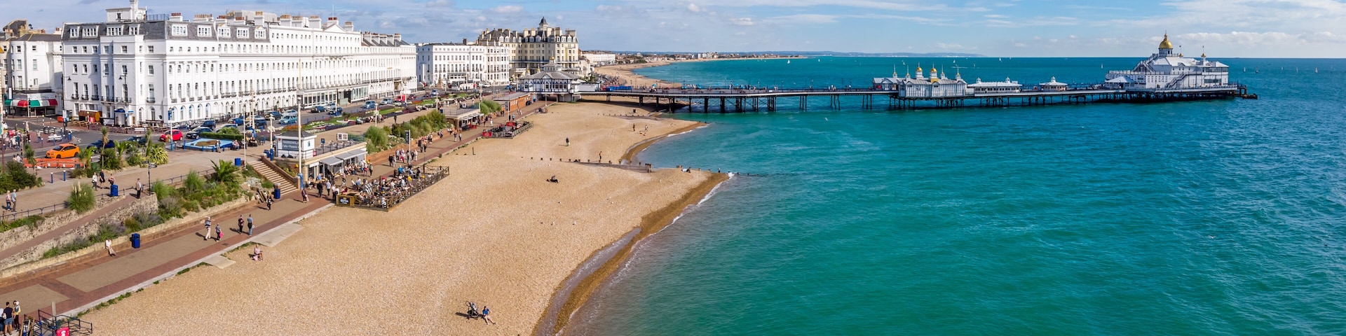Un paisaje urbano costero con una playa arenosa, un muelle que se adentra en el mar y edificios históricos a lo largo de la costa.