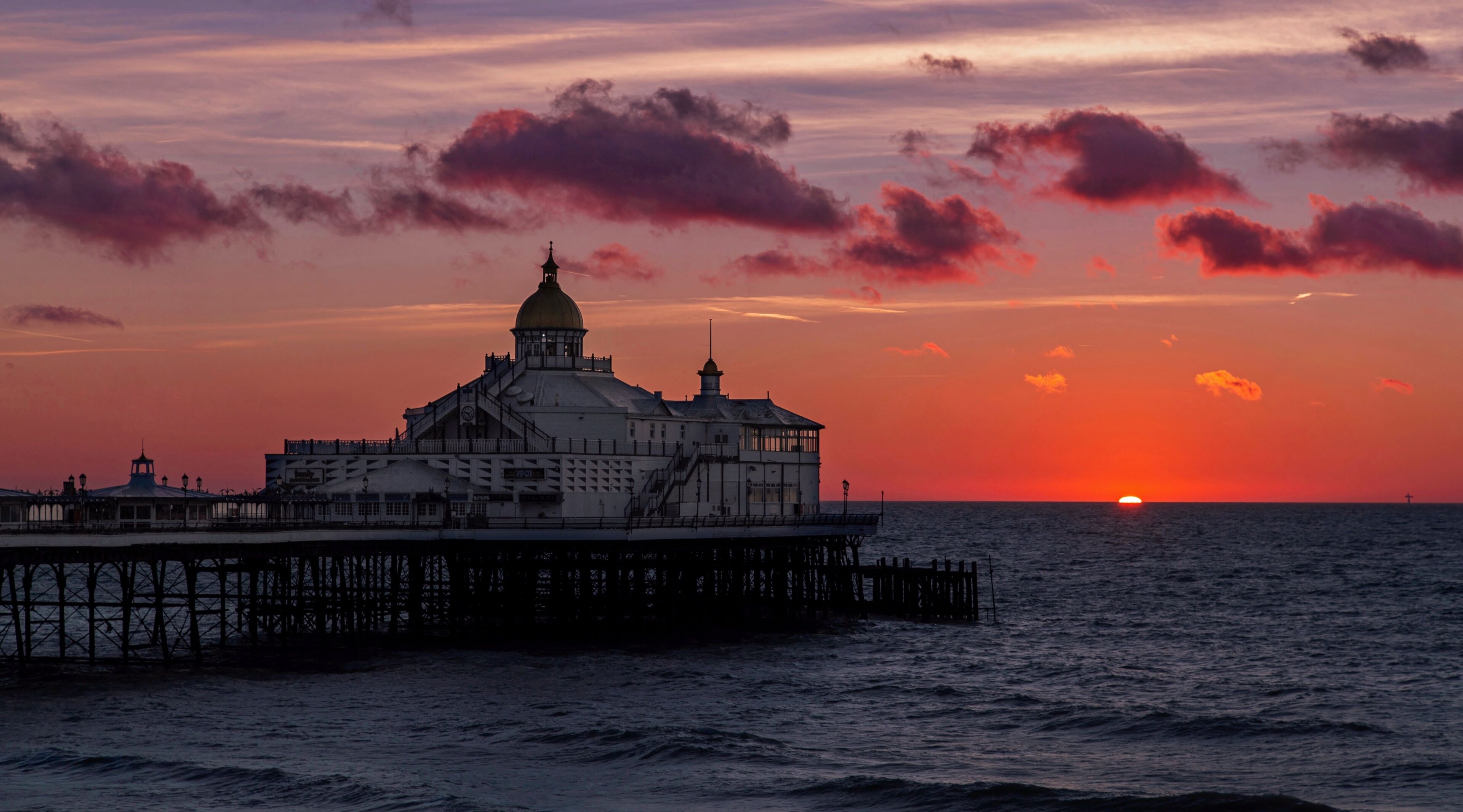 A coastal pier with a prominent building featuring a dome and a clock tower, set against a sunset with clouds.