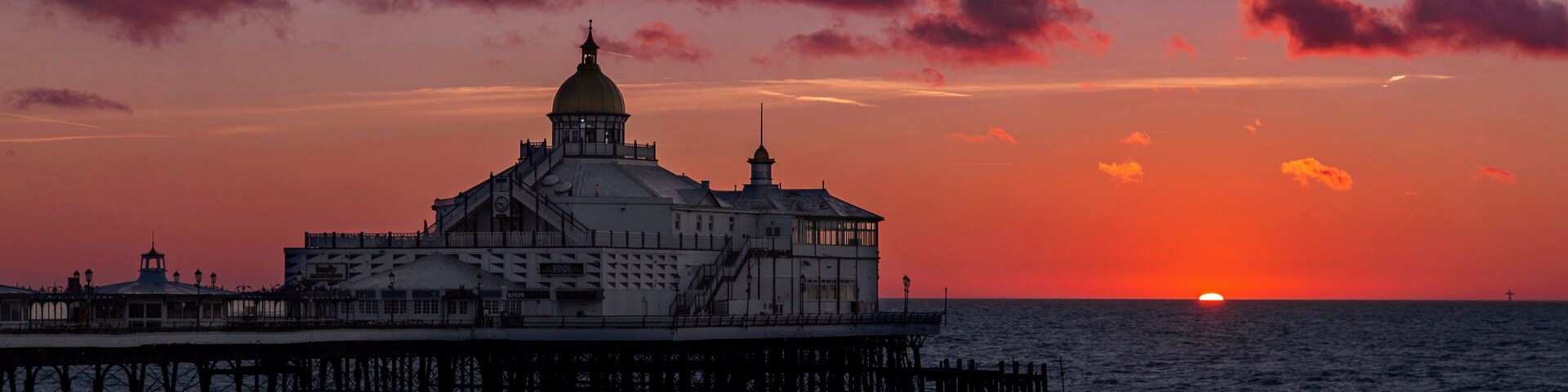 Um píer costeiro com um edifício imponente, com cúpula e torre do relógio, ao pôr do sol com nuvens.