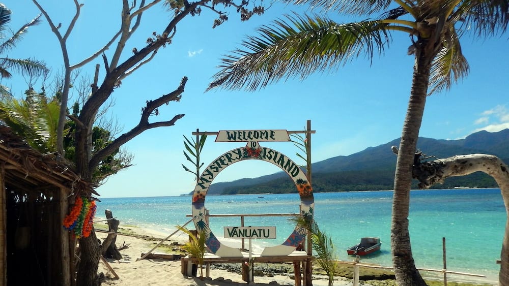 A beach with a 'Welcome to Vanuatu' sign, palm trees, and clear blue water.