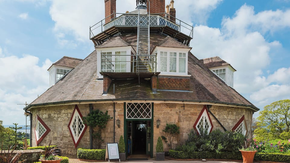 A large, historic building with a prominent tower, stone walls, and a well-maintained garden.