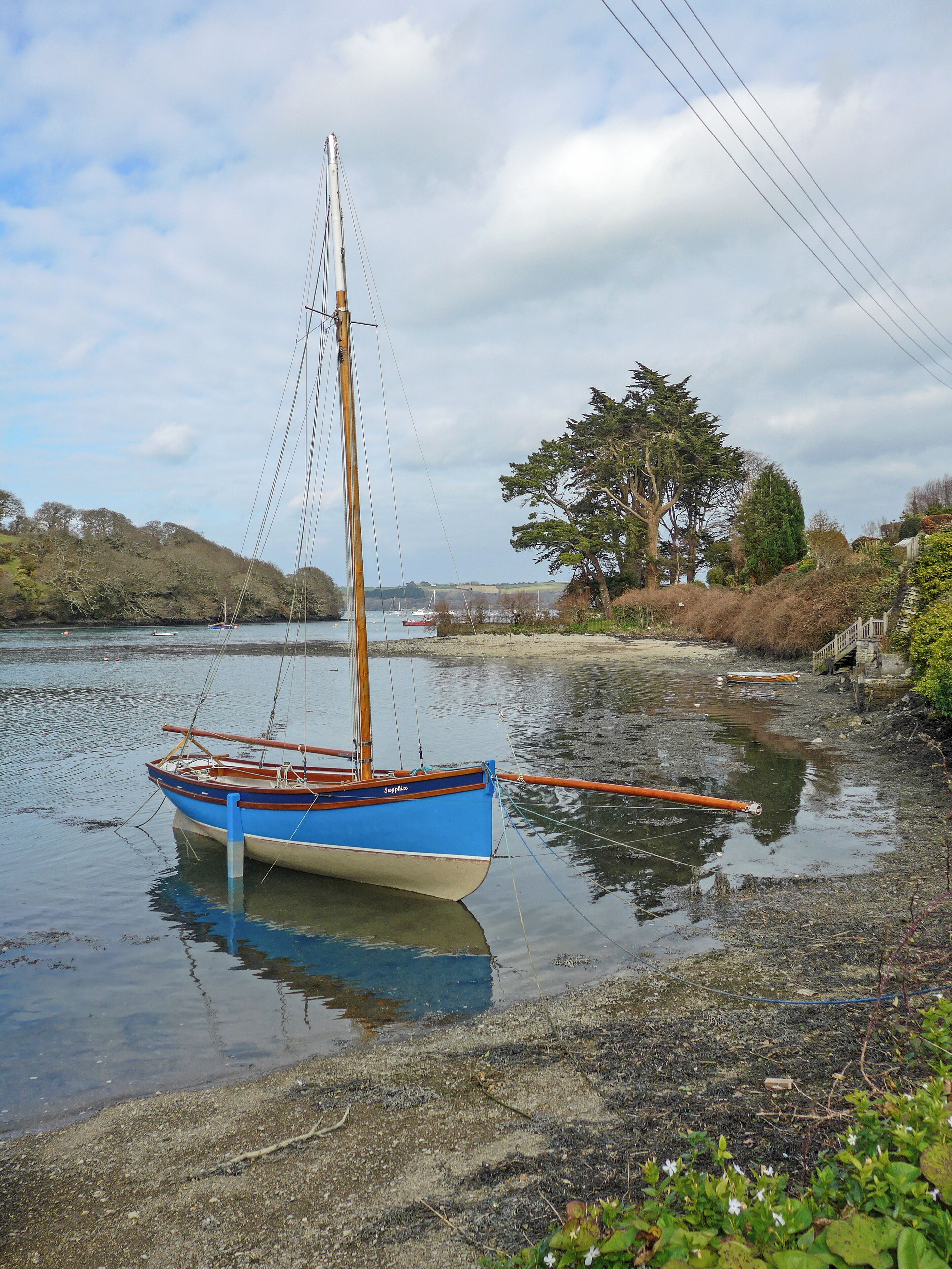 A blue boat named 'Sapphique' docked at a calm riverbank with trees and a wooden bridge in the background.