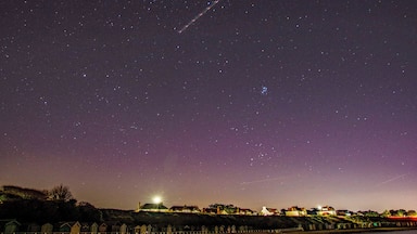 Céu estrelado sobre uma cidade costeira, com um rastro visível de condensação.