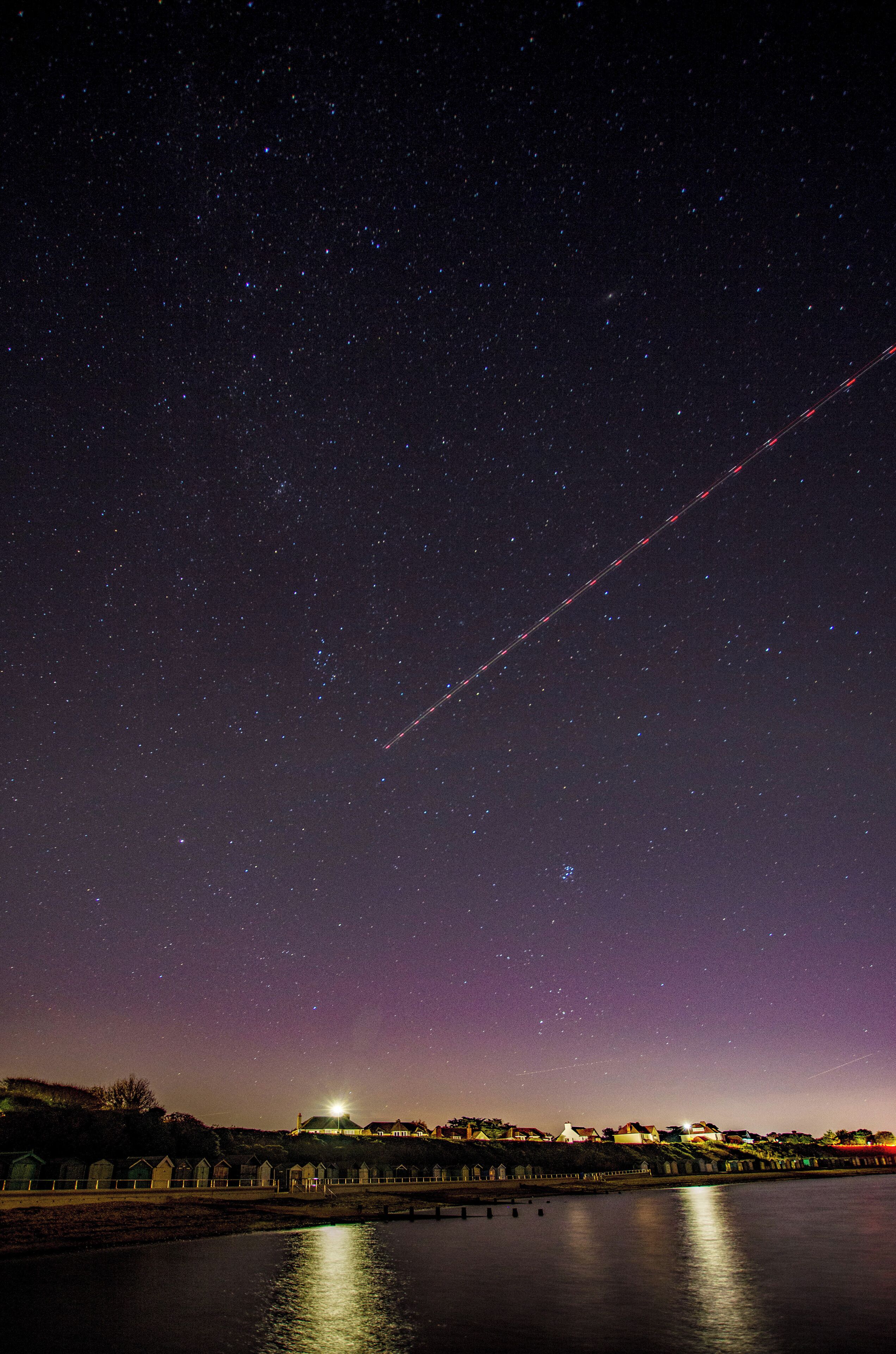 A starry night sky over a coastal town with a visible contrail.