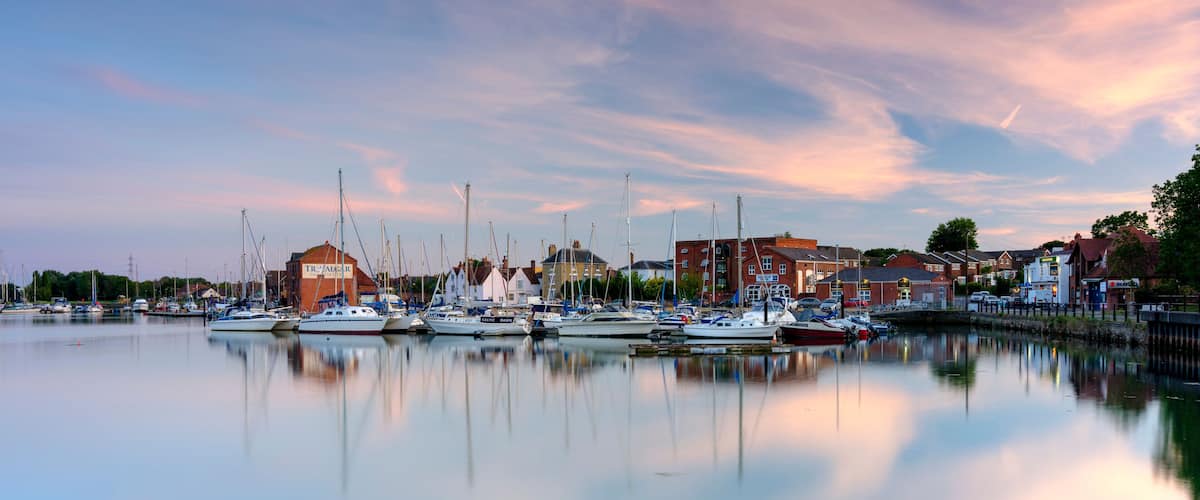 A marina with boats docked, buildings, and a calm water body reflecting the sky.