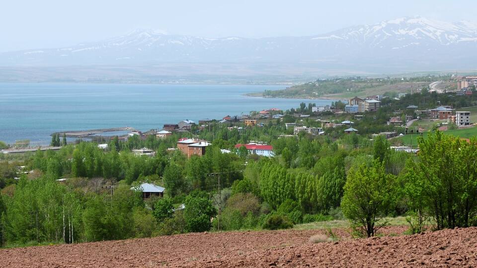 A coastal town with a lake, green hills, and snow-capped mountains in the background.
