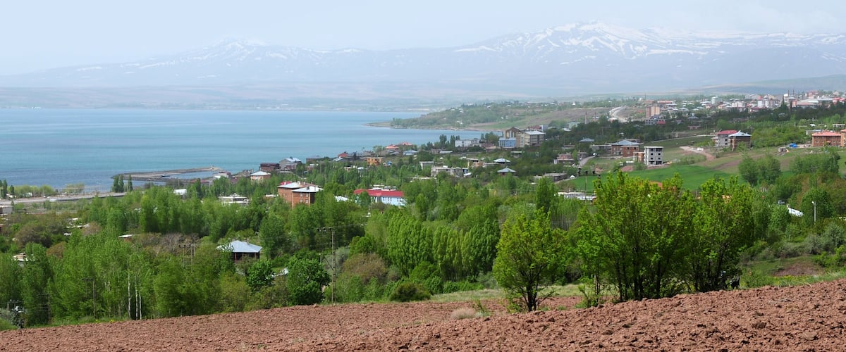 A coastal town with a lake, green hills, and snow-capped mountains in the background.