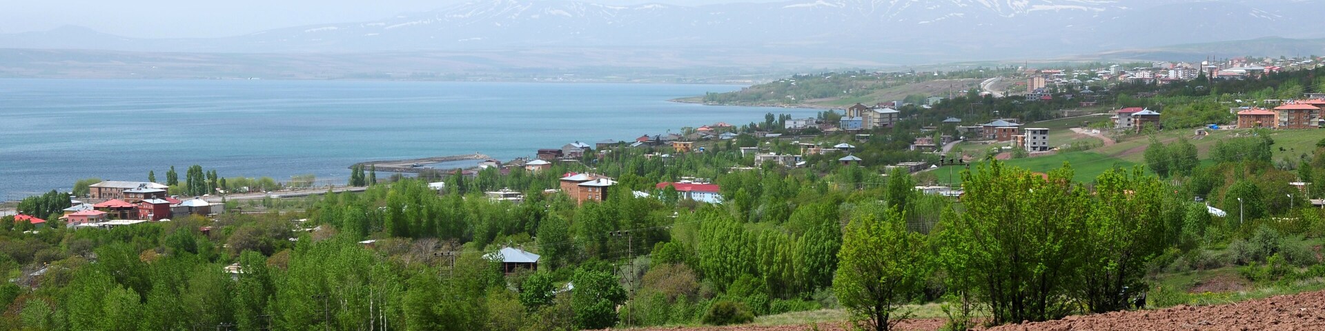 A coastal town with a lake, green hills, and snow-capped mountains in the background.