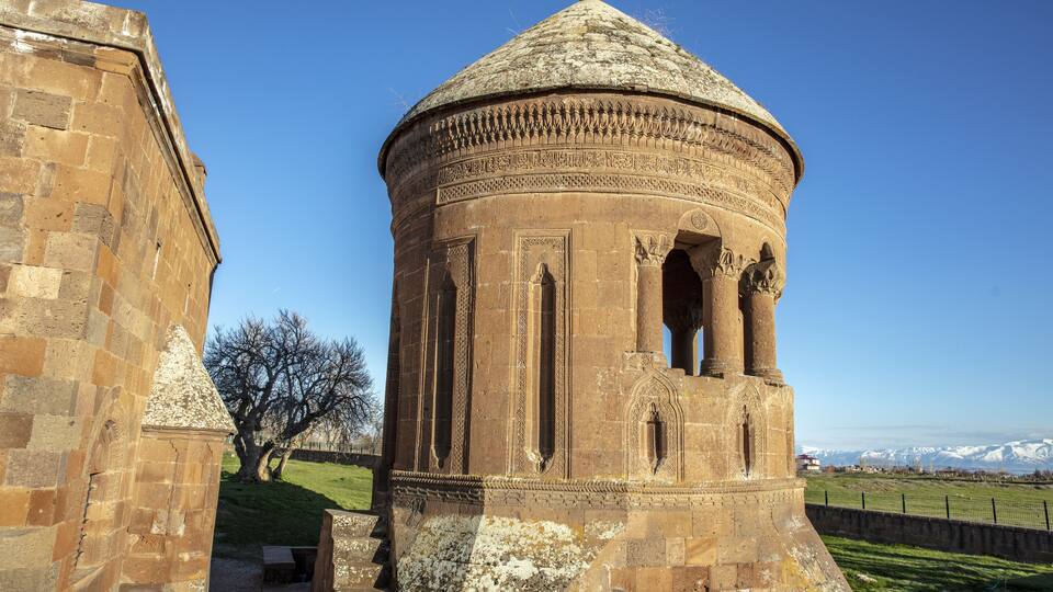 A historic stone tower with intricate carvings and a dome, surrounded by a grassy field and a clear blue sky.